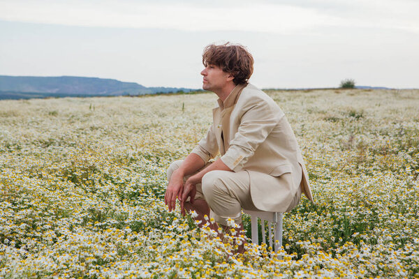 Tall handsome man dressed in a white suit on naked body sitting on a back of a chair in camomile flowers field