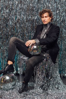 Uzun boylu çekici bir adam ışıltılı arka planda sandalyede oturuyor ve elinde ayna disko topu var. Parti konsepti.