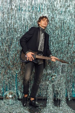 Parlak arka planda elektro gitar tutan uzun boylu çekici bir adam ve yerde ayna disko topu var. Parti konsepti.