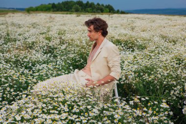 Beyaz takım elbiseli uzun boylu yakışıklı adam papatya çiçeği tarlasında beyaz bir sandalyede oturuyor.