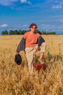 Kırsal Kır Sahnesi. Siyah gömlek, siyah şapka ve pembe güneş gözlüğü takmış uzun boylu yakışıklı bir adam altın yulaf tarlasında tek bacaklı kahverengi deri bir valizin üzerinde duruyordu. Mavi gökyüzü ile yaz manzarası