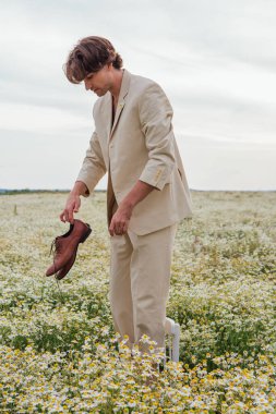 Uzun boylu, beyaz takım elbiseli, çıplak vücutlu, papatya çiçekli sandalyede çıplak ayakla duran ve elinde ayakkabıları olan yakışıklı bir adam.
