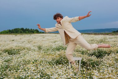 Uzun boylu, beyaz takım elbiseli, çıplak vücutlu, papatya çiçeği tarlasında beyaz sandalyede dans eden yakışıklı bir adam.