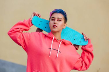 Omuzlarında mavi plastik kaykay tutan kısa saçlı şık genç bir kadın. Gençlik kavramı.