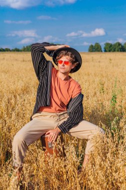 Kırsal Kır Sahnesi. Siyah gömlekli, siyah şapkalı ve pembe gözlükle altın yulaf tarlasında kahverengi deri bir valizde oturan uzun boylu yakışıklı bir adam. Mavi gökyüzü ile yaz manzarası