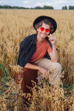 Kırsal Kır Sahnesi. Siyah gömlekli, siyah şapkalı ve pembe güneş gözlüklü uzun boylu yakışıklı bir adam altın yulaf tarlasında oturmuş, içi yulaf dolu kahverengi deri valiziyle..
