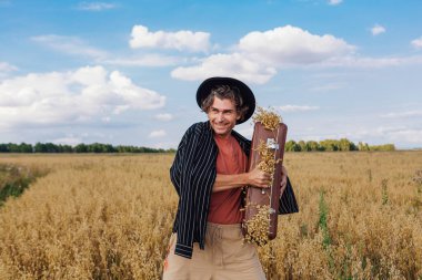 Kırsal Kır Sahnesi. Siyah gömlekli ve siyah şapkalı uzun boylu yakışıklı adam altın yulaf tarlasında kulaklarla dolu kahverengi klasik deri bavula sarılıyor.