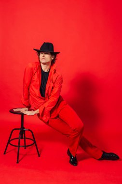 Kırmızı gömlekli, uzun boylu, yakışıklı bir adamın portresi. Kırmızı pantolonlu ve siyah şapkalı. Kırmızı arka planda sandalyeli.