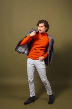 Turuncu balıkçı yaka, gri ceket ve yeşil arka planda beyaz kot giyen uzun boylu yakışıklı bir adamın tam portresi.