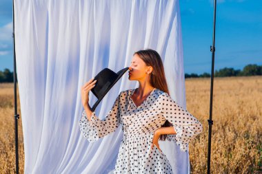 Kırsal Kır Sahnesi. Beyaz benekli elbiseli, siyah şapkalı, uzun saçlı, altın yulaf tarlasında beyaz perdeli genç bir kadın..