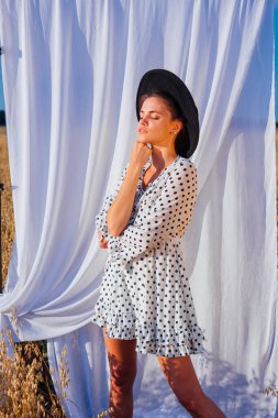 Kırsal Kır Sahnesi. Beyaz benekli elbiseli, siyah şapkalı, uzun saçlı, altın yulaf tarlasında beyaz perdeli genç bir kadın..