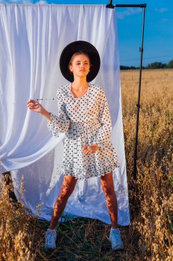 Kırsal Kır Sahnesi. Beyaz benekli elbiseli, siyah şapkalı, uzun saçlı, altın yulaf tarlasında beyaz perdeli genç bir kadın..