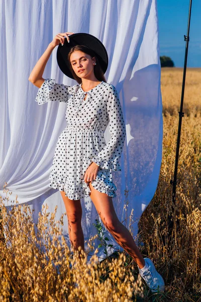 Kırsal Kır Sahnesi. Beyaz benekli elbiseli, siyah şapkalı, uzun saçlı, altın yulaf tarlasında beyaz perdeli genç bir kadın..