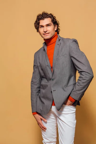 Tall handsome man dressed in orange turtleneck, white jeans and grey jacket posing on the beige background