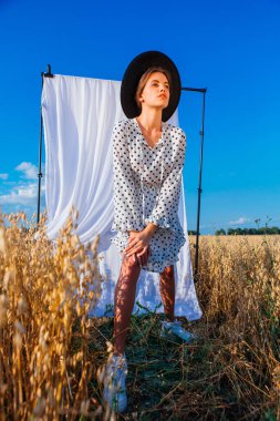 Kırsal Kır Sahnesi. Beyaz benekli elbiseli, siyah şapkalı, uzun saçlı, altın yulaf tarlasında beyaz perdeli genç bir kadın..