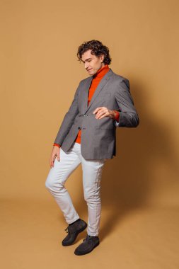 Turuncu balıkçı yaka, beyaz kot pantolon ve bej arka planda duran gri ceketli uzun boylu yakışıklı bir adamın moda portresi.
