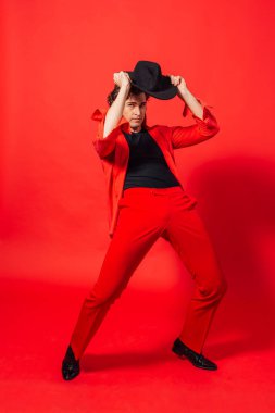 Kırmızı gömlekli, siyah şapkalı, kırmızı arka planda poz veren uzun boylu yakışıklı bir adamın portresi..