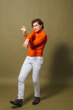 Turuncu balıkçı yaka ve yeşil arka planda duran beyaz kot giyen uzun boylu yakışıklı bir adamın tam portresi.