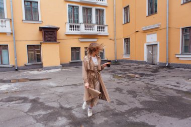 Genç gülümseyen milenyum kadını, yaban saçlarıyla sonbahar paltosu giymiş elinde bir fincan kahveyle eski binanın yakınına gidiyor..