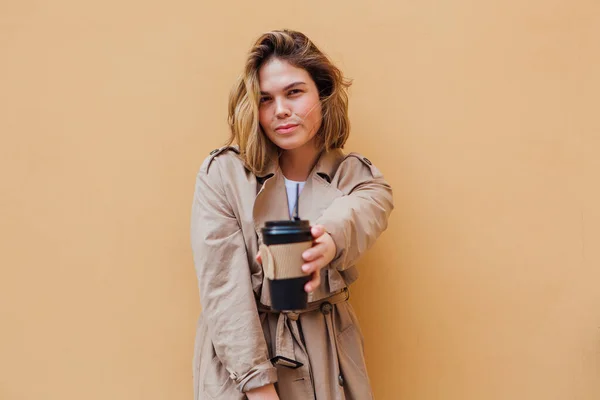 Young smiling millennial woman with wild hair dressed in an autumn coat standing a cup of coffee to go near the beige wall.