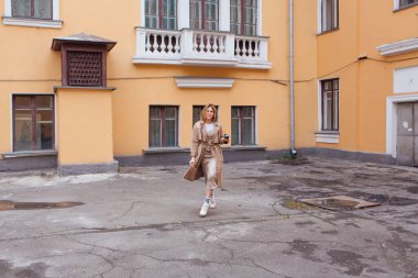 Genç gülümseyen milenyum kadını, yaban saçlarıyla sonbahar paltosu giymiş elinde bir fincan kahveyle eski binanın yakınına gidiyor..