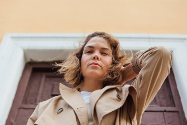 Genç milenyum kadını, eski bir binanın kapısında poz veren, sonbahar paltosu giymiş vahşi saçlı..