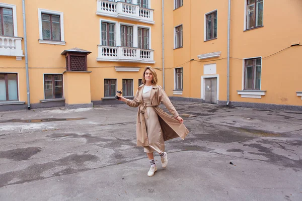 Joven mujer milenaria con pelo salvaje vestida con un abrigo de otoño caminando con una taza de café para acercarse al viejo edificio. — Foto de Stock