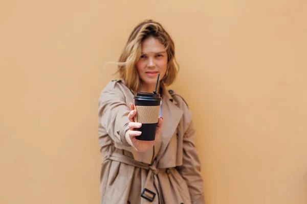 Young smiling millennial woman with wild hair dressed in an autumn coat standing a cup of coffee to go near the beige wall.