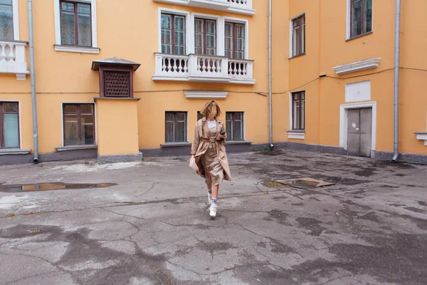 Young smiling millennial woman with wild hair dressed in an autumn coat walking with a cup of coffee to go near the old building.