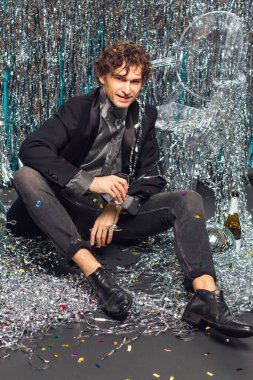 Uzun boylu çekici bir adam, ışıltılı arka planda yerde oturuyor ve ayna disko topu elinde şampanya kadehi tutuyor. Parti konsepti.