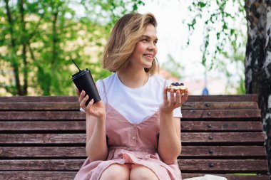 Parktaki bankta oturan genç romantik kadın güneşli bir yaz gününde yanında kek ve bir fincan kahveyle