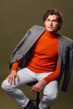 Turuncu balıkçı yaka, gri ceket ve yeşil arka planda beyaz kot giyen uzun boylu yakışıklı bir adamın tam portresi.