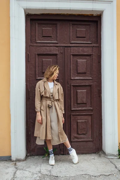 Young smiling millennial woman with wild hair dressed in an autumn coat posing near the door of an old building.