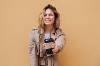 Genç gülümseyen milenyum kadını, sonbahar montu giymiş, bej duvarın yanına gitmek için bir fincan kahve taşıyan vahşi saçlı..