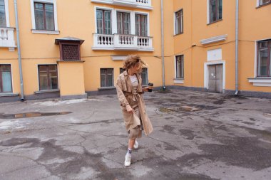 Genç milenyum kadını, sonbahar paltosu giymiş vahşi saçlarıyla elinde bir fincan kahveyle eski binanın yakınına gidiyor..