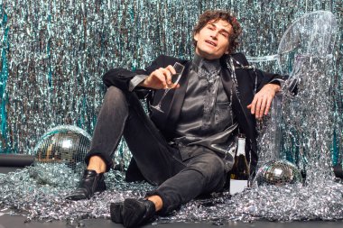 Uzun boylu çekici bir adam, ışıltılı arka planda yerde oturuyor ve ayna disko topu elinde şampanya kadehi tutuyor. Parti konsepti.