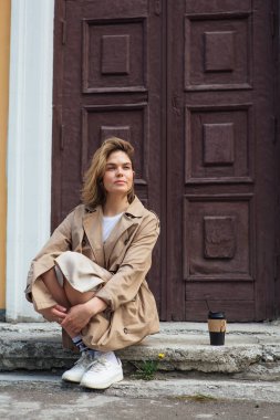 Genç gülümseyen milenyum kadını, sonbahar paltosu giymiş vahşi saçlı, elinde bir fincan kahveyle eski bir binanın kapısında oturuyor..