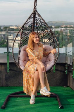Uzun leopar elbiseli güzel bir kadın bir binanın çatısında salıncakta oturuyor..