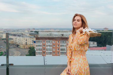 Uzun leopar desenli elbisesi içinde güzel bir kadın binanın çatısında elinde beyaz ayakkabılarıyla merdivenlerde dikiliyor..