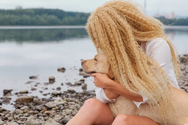 閉じる上のポートレート若いです美しいです女性とともにブロンド巻き毛ハグとともに彼女のラブラドール取得犬オンザ川の海岸 — ストック写真