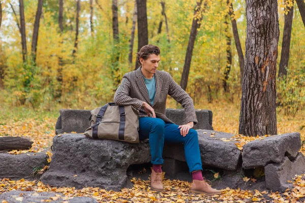 背の高いハンサムな男茶色のジャケットを身に着けている秋の森の岩の上に袋でポーズ — ストック写真
