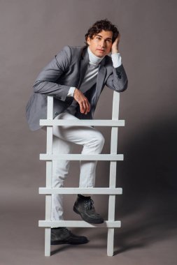 Beyaz balıkçı yaka, beyaz kot pantolon ve gri arka planda beyaz merdivenle poz veren uzun boylu yakışıklı bir adamın moda portresi.