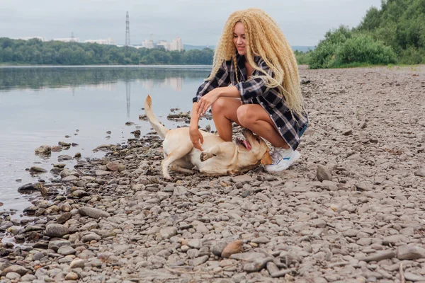 若いです美しいです女性とともにブロンド巻き毛あります遊びますともに彼女のラブラドール取得犬オンザ川の海岸 — ストック写真