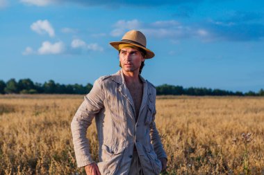 Kırsal kesimde. Altın yulaf tarlasında duran kalın keten takım elbiseli ve şapkalı uzun boylu yakışıklı bir adam.
