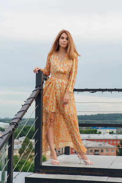 Une fille en robe longue debout avec les pieds nus sur les escaliers sur le toit du bâtiment. — Photo