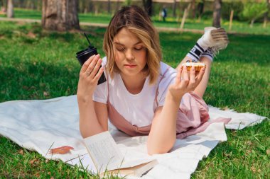 Romantik genç bir kadın açık bir kitap, kek ve bir fincan kahveyle parkta çimlere uzanıyor güneşli bir yaz gününde.