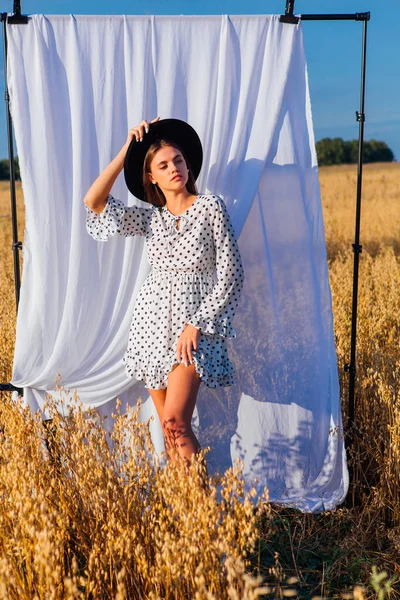 Kırsal Kır Sahnesi. Beyaz benekli elbiseli, siyah şapkalı, uzun saçlı, altın yulaf tarlasında beyaz perdeli genç bir kadın..