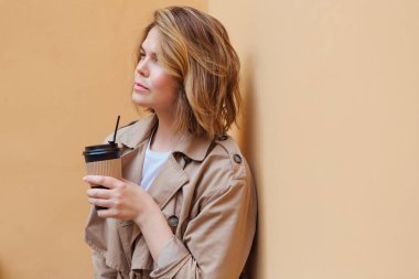 Genç gülümseyen milenyum kadını, sonbahar montu giymiş, bej duvarın yanına gitmek için bir fincan kahve taşıyan vahşi saçlı..