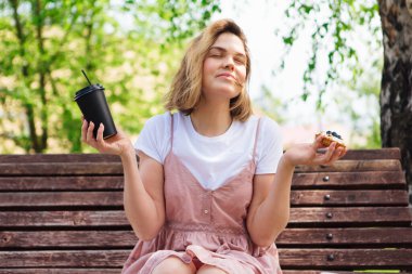 Parktaki bankta oturan genç romantik kadın güneşli bir yaz gününde yanında kek ve bir fincan kahveyle