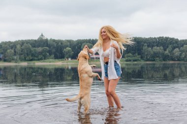 Sarışın, kıvırcık saçlı, labrador köpeğiyle nehirde oynayan genç ve güzel bir kadın..
